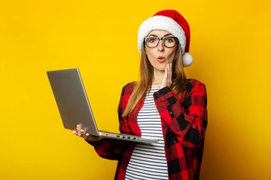 Sarı arka planda dizüstü bilgisayarı tutan, Noel Baba şapkalı ve kırmızı kareli tişörtlü, şaşkın yüzlü genç bir kadın..