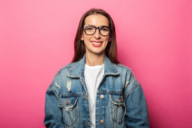 Gözlüklü, pembe arka planda kot ceketli, arkadaş canlısı genç bir kadın.