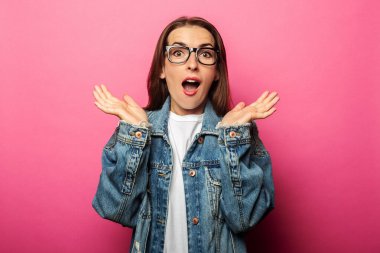 Pembe arka planda gözlüklü ve kot ceketli genç bir kadın.