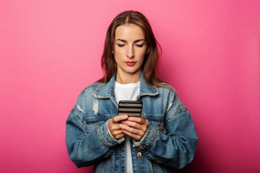 Kot ceketli genç kız pembe arka plandaki telefona bakıyor.