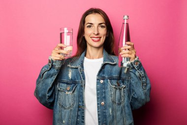 Gülümseyen genç bir kadın pembe arka planda su şişesi ve bir bardak su tutuyor.