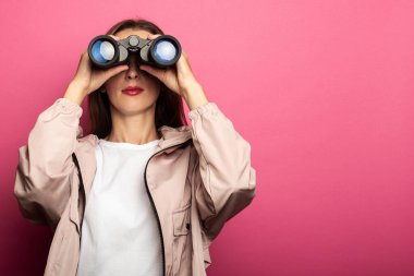 Ceketli genç bir kadın pembe arka planda dürbünle bakıyor.