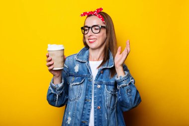 Kot ceketli, bantlı ve gözlüklü genç kadın sarı arka plandaki kağıt bardağa şaşkın bakıyor..