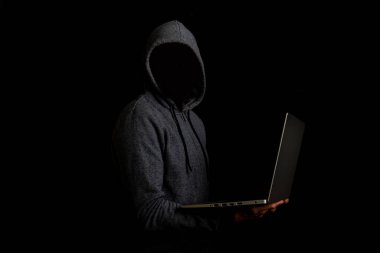 Kapüşonlu yüzü olmayan bir adam karanlık bir arka planda dizüstü bilgisayar tutuyor. Siber dolandırıcı kavramı, hacker. Pankart.