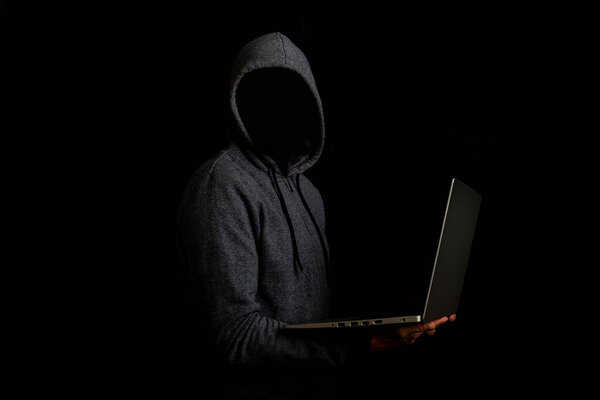 Человек без лица в капюшоне держит ноутбук на тёмном фоне. Концепция кибер-мошенника, хакер. Баннер.