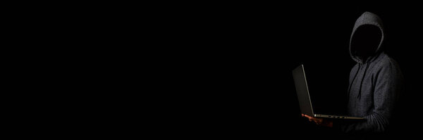 Человек без лица в капюшоне держит ноутбук на тёмном фоне. Концепция кибер-мошенника, хакер. Баннер.