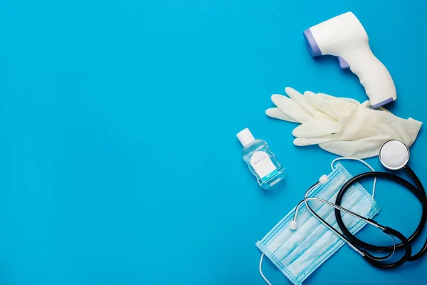Digital non-contact thermometer, protective mask, latex gloves, medical stethoscope and sanitizer on a blue background. Banner. Top view, flat lay.