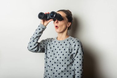 Süveter giymiş, şaşırmış yüzlü genç bir kadın arka plandaki dürbünle bakar..