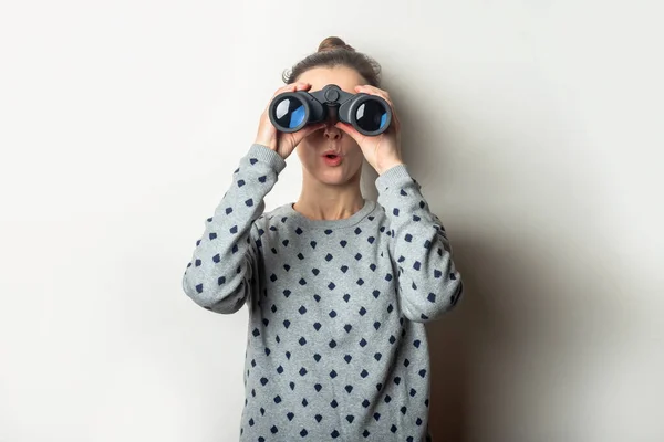 Young woman with a surprised face in a sweater looks through binoculars on a light background.