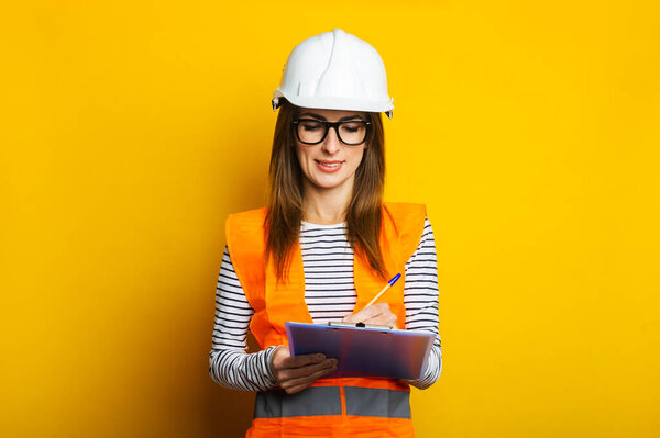 Young woman in a vest and hard hat holds a clipboard and takes notes on a yellow background. Concept for construction, new building, renovation. Banner.