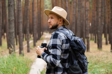 Sırt çantası ve çam ormanında dürbünlü şapkalı genç adam. Dağlarda ya da ormanlarda yürüyüş.