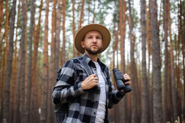 Sırt çantası ve çam ormanında dürbünlü şapkalı genç adam. Dağlarda ya da ormanlarda yürüyüş.