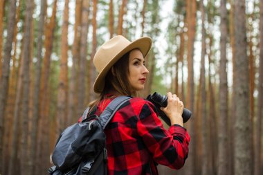Sırt çantası, şapka ve kırmızı ekose gömlekli genç bir turist ormanda dürbün tutuyor..
