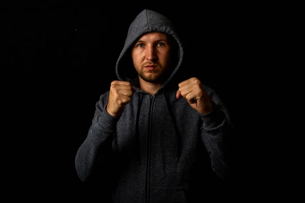 Aggressive young bearded man in a sweatshirt in a hood shows his fists on a black background