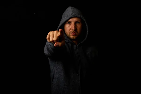 Man in a hood points a finger against a dark background.