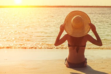 Gün batımında genç bir kız plajda oturuyor. Sırtı denize dönük bir şapkayla.