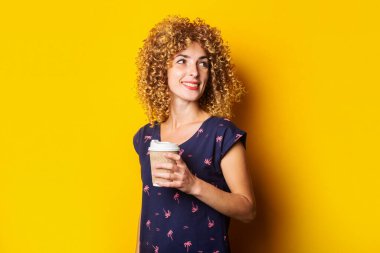 Sarı arka planda kağıt bardak tutan kıvırcık saçlı, gülümseyen genç kız.