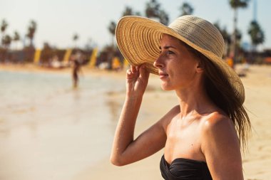 Kumsalda oturan genç kız şapkasını tutar ve denize bakar.