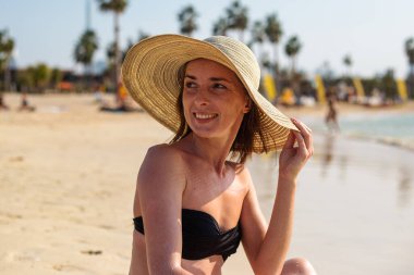 Kumsalda oturmuş şapkasıyla gülümseyen genç bir kız.