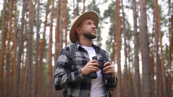 Un homme barbu et un chapeau avec des jumelles marchant dans les bois. Composition randonnée, forestier. — Video