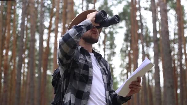 El hombre con sombrero sostiene un mapa y mira a través de los prismáticos mientras camina por el bosque. Concepto de orientación, viajes. — Vídeo de stock