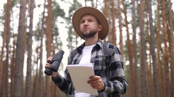 El hombre con sombrero sostiene un mapa y mira a través de los prismáticos mientras camina por el bosque. Concepto de orientación, viajes. — Vídeo de stock