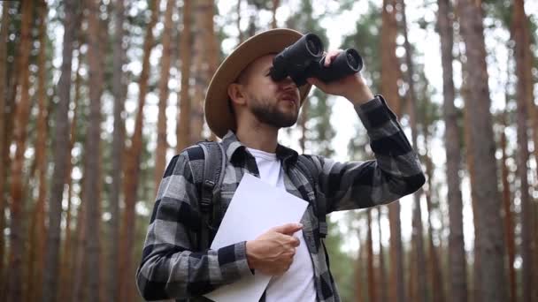 El hombre con sombrero sostiene un mapa y mira a través de los prismáticos mientras camina por el bosque. Concepto de orientación, viajes. — Vídeo de stock