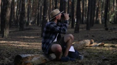 Genç adam bir kütüğün üzerinde dinleniyor ve yürürken dürbünle bakıyor.