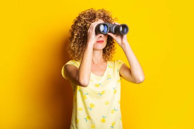 Kıvırcık genç kadın parlak sarı bir arka planda dürbünle bakar.