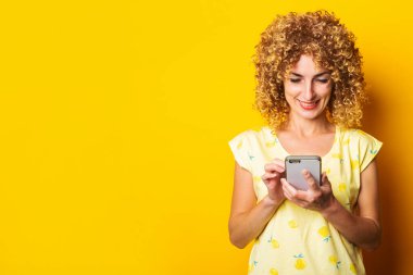 Kıvırcık, gülümseyen genç bir kadın sarı arka plandaki telefona bakar.