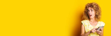 Kıvırcık genç kadın sarı arka plandaki telefonu işaret ederken şaşırdı. Pankart