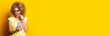 Sarı arka plandaki telefona büyüteçle bakan, şok olmuş kıvırcık bir genç kadın. Pankart