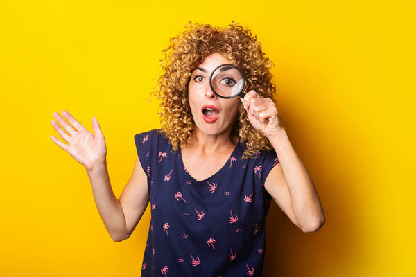surprised curly young woman looks at the camera through a magnifying glass on a yellow background