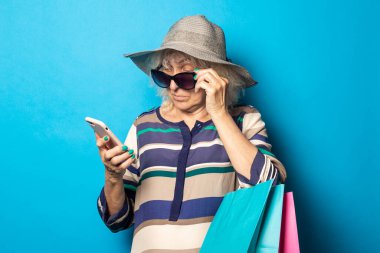 Şapkalı ve gözlüklü yaşlı kadın alışveriş çantalarını tutuyor ve mavi arka planda telefonuna bakıyor..