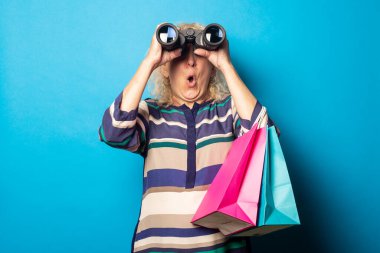 Şaşkın suratlı, alışveriş torbaları tutan ve mavi arka plandaki dürbünlerden bakan yaşlı kadın..