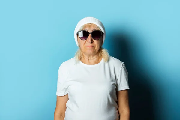 Funny old woman in a hat and glasses, makes a hand gesture on a blue background. Concept cool stylish grandmother, modern style.