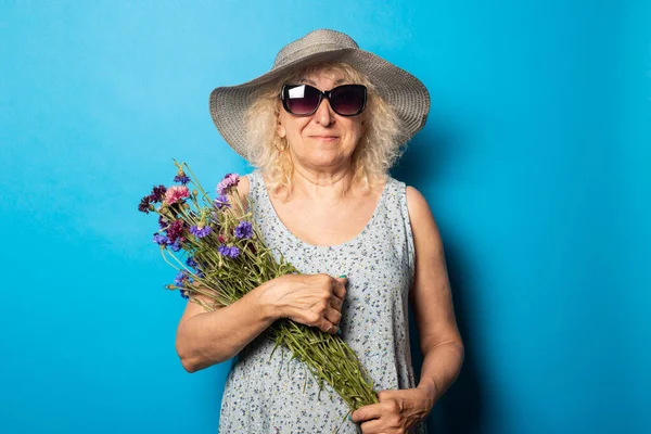 Old woman with a smile in a hat with wide brim and a dress holding a bouquet of flowers on a blue background.