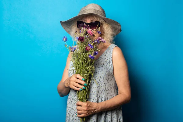 Old woman in a wide-brimmed hat and dress sniffing a bouquet of flowers on a blue background.
