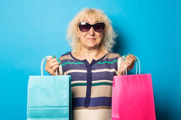 Smiling old woman holding shopping bags on blue background.