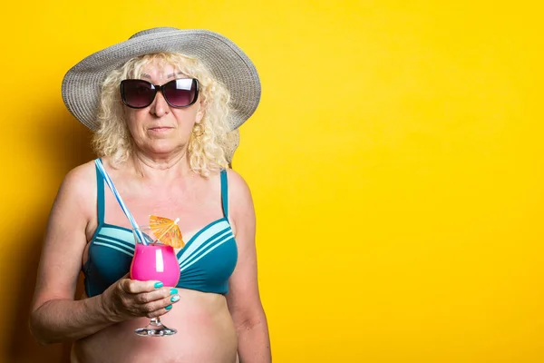 Old woman in swimsuit wearing hat and sunglasses holding cocktail on yellow background