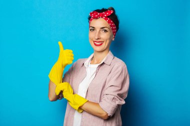 Gülümseyen genç kadın mavi arka planda temizlik eldivenleriyle baş parmağını kaldırıyor.