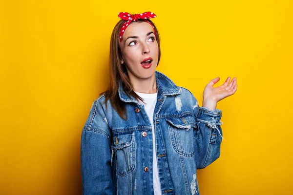 Young woman in a hair band, in a denim jacket, looks surprised to the side on a yellow background