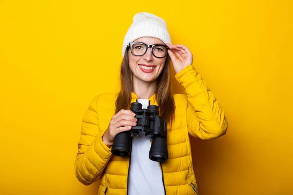 Smiling young woman in yellow jacket holding binoculars on yellow background