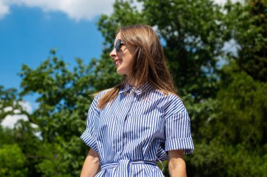 Parkta gözlüklü ve elbiseli genç bir kadın duruyor..