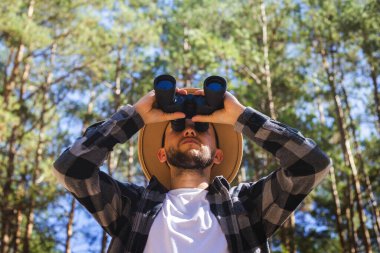 Şapkalı ve gri ekose gömlekli bir turist dürbünle bakar..