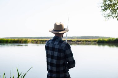 Şapkalı ve ekose gömlekli bir adam nehir kıyısında balık tutuyor..