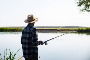 Şapkalı ve ekose gömlekli bir adam nehir kıyısında balık tutuyor..