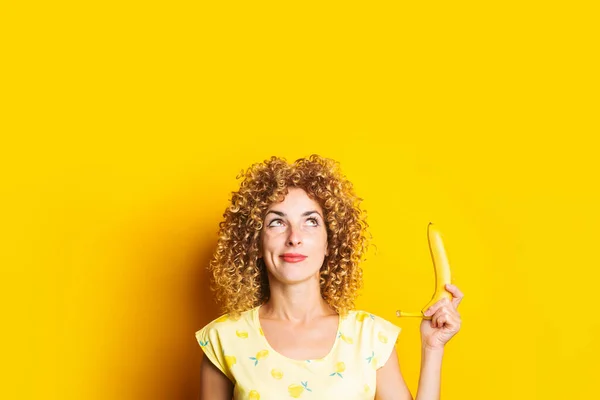 Curly Young Woman Holding Banana Pensively Looking Upwards Yellow Background — Stock Photo, Image