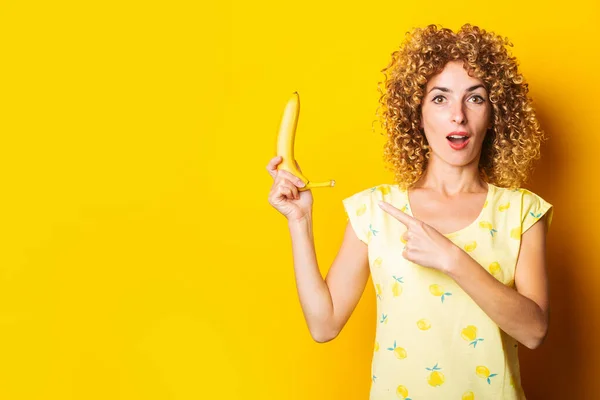 Шокированная Кудрявая Молодая Женщина Держит Банан Указывая Пальцем Желтый Фон — стоковое фото