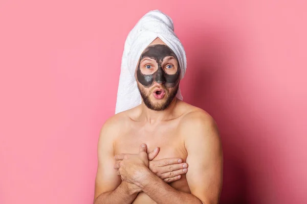 nude young man with a cosmetic mask on his face, hiding his chest with his hands on a pink background.